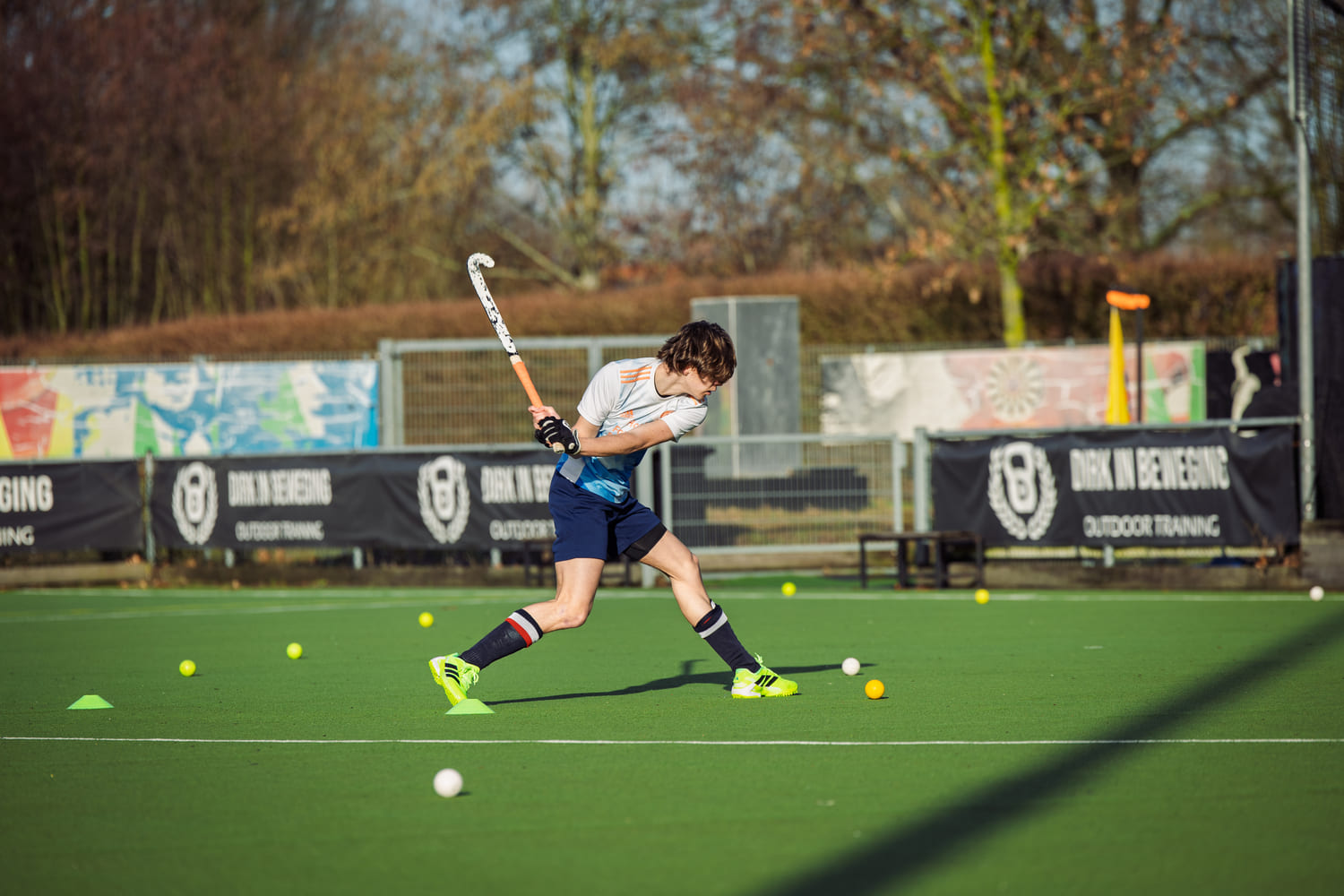 Hockeyspeler in actie tijdens ProHockey training op groen veld met felgroene schoenen en witte en gele ballen op de achtergrond.