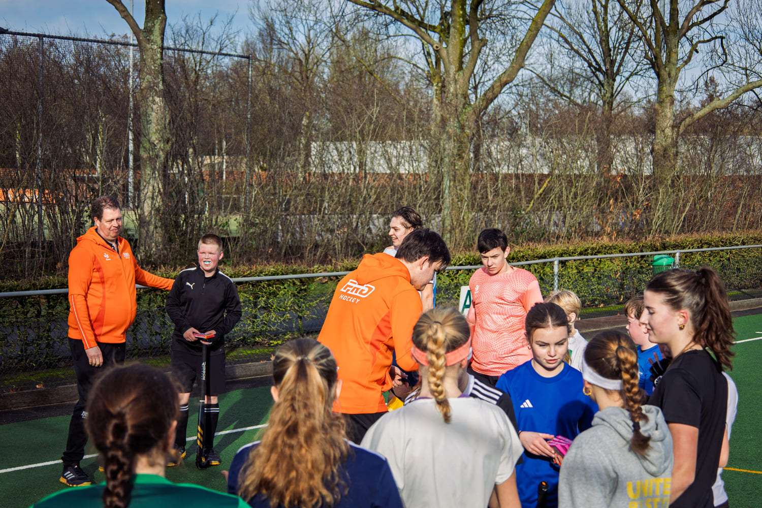 Groep jonge hockeyspelers en trainers verzamelen zich op een veld tijdens een ProHockey-kamp, gekleed in sportieve oranje en blauwe outfits.