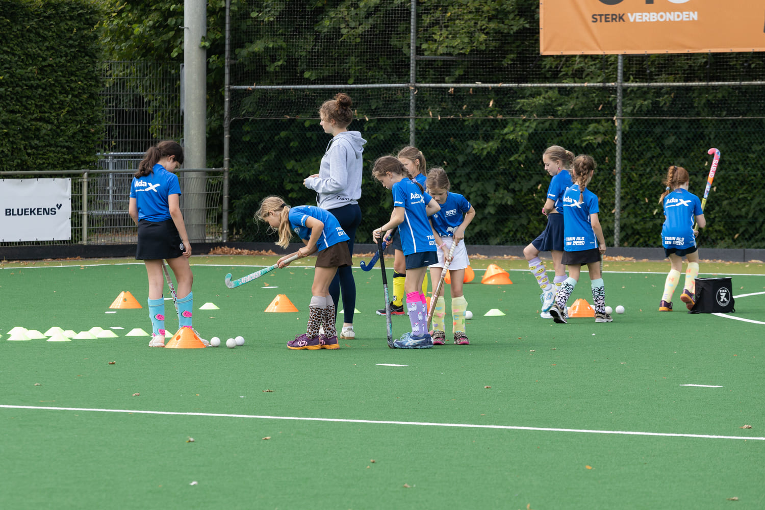 Kinderen oefenen hockey bij ProHockey zomerkamp op het veld van hockeyclub Prinsenbeek, begeleid door een coach.